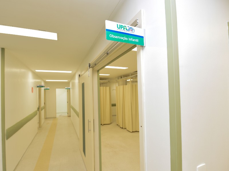 Sala de observação infantil da UPA Tatuapé com paredes brancas. Do lado direito há uma porta ampla com uma placa escrito UPA 24 horas Observação Infantil. Ao fundo ficam os leitos separados por cortinas em tom de bege